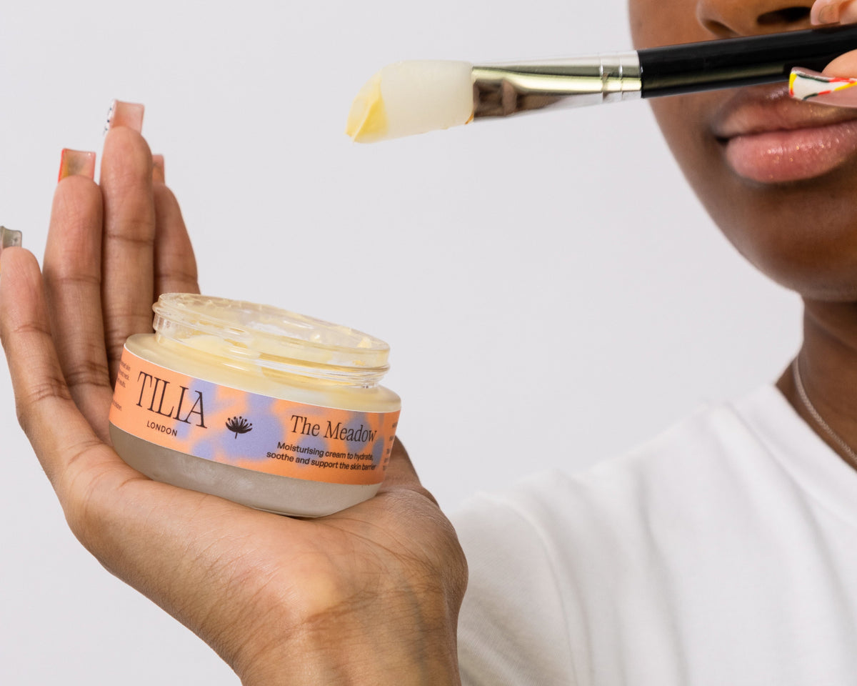A close-up of a person holding an open jar of Tilia London 'The Meadow' moisturizing cream while applying it with a makeup brush. The person is wearing a white t-shirt against a clean white background.